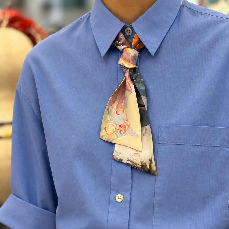 A close-up of a person wearing a light blue button-up shirt with rolled sleeves, a colorful abstract-patterned necktie, and brown trousers.