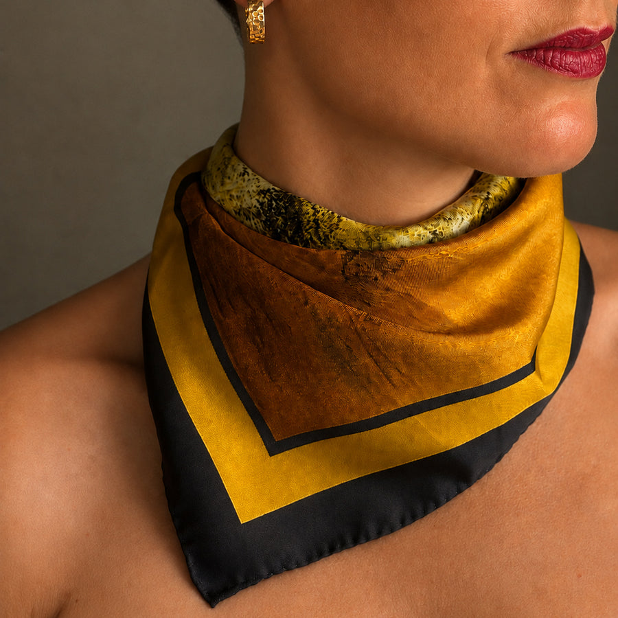 A close-up of a woman wearing a square silk scarf styled as a collar. The scarf features a textured mustard yellow top section, a wide black border, and a yellow and black abstract patterned inner section.