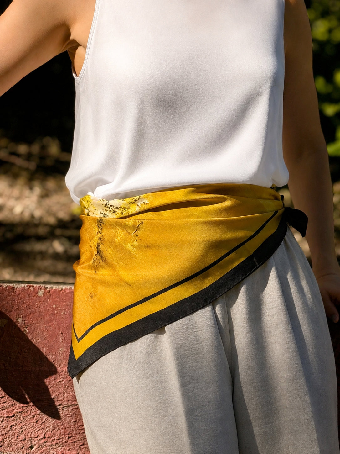 A mustard yellow wrap-style belt with black trim and a small white floral accent, worn around the waist of a person in a white sleeveless top and light gray pants.