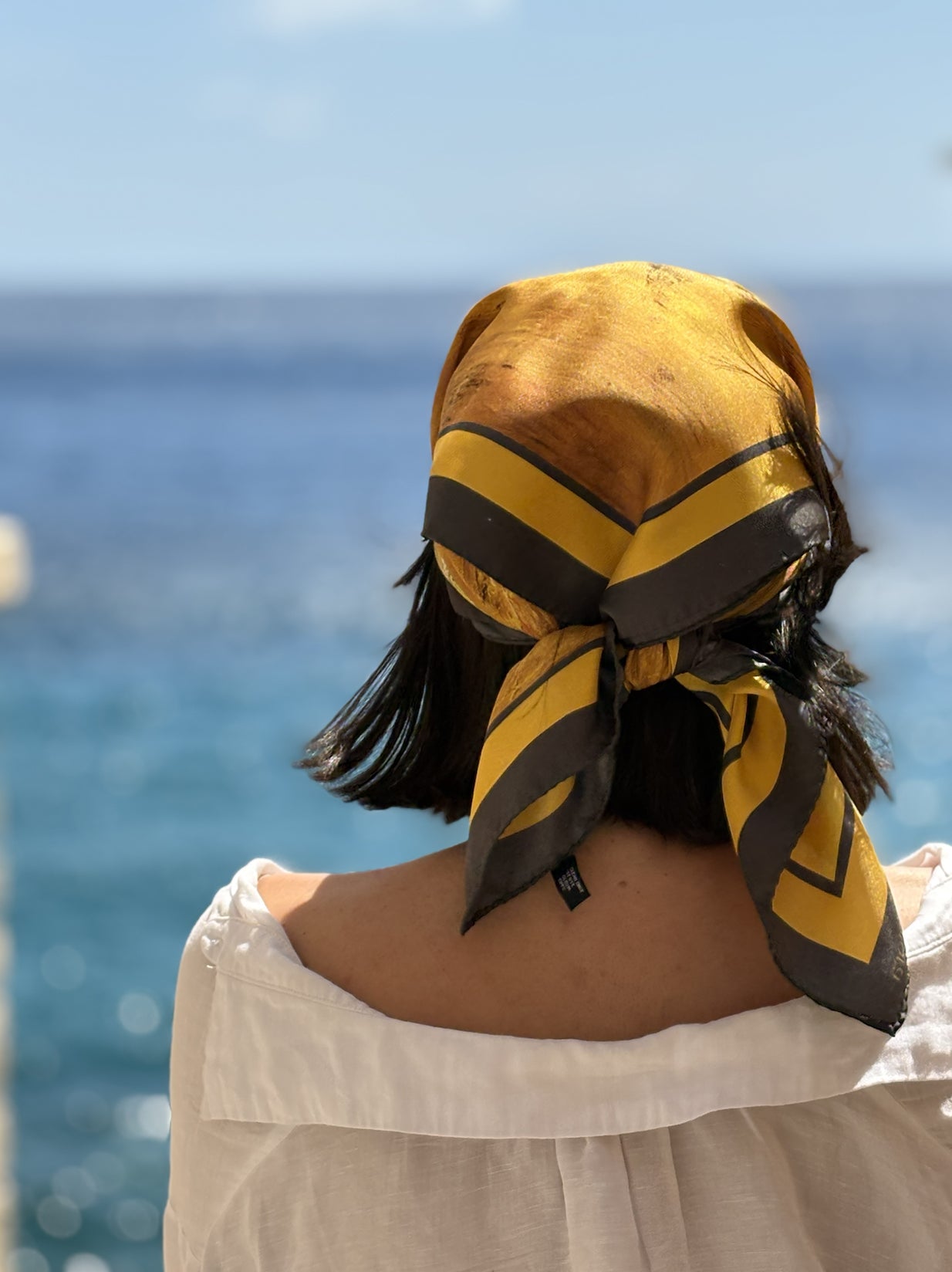 A person seen from behind wearing a yellow and black striped silk scarf tied around their head, with a blurred blue ocean and sky background.