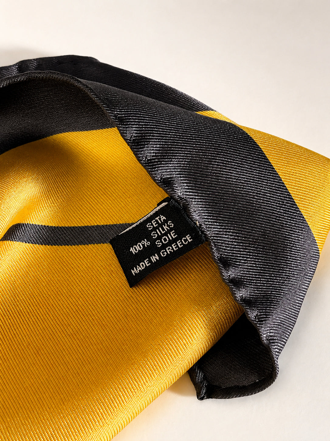 Close-up of a yellow and black striped silk scarf with a black fabric tag showing 'SETA SILKS S.O.E. MADE IN GREECE'.