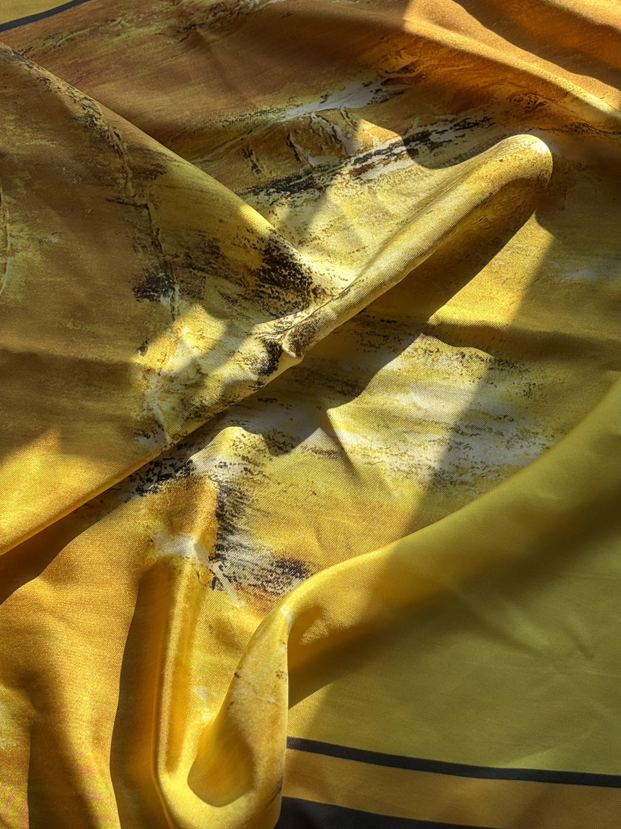 A close-up of a draped yellow fabric with a subtle abstract black and white pattern, showing soft folds and shadows under warm light.