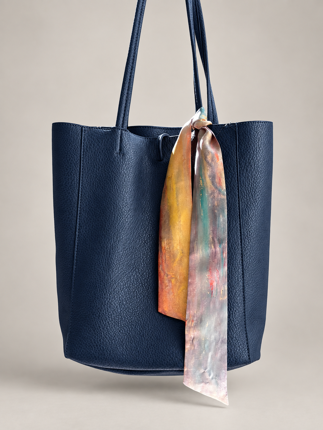 A navy blue pebbled leather tote bag with dual shoulder straps, featuring a colorful silk scarf tied to the side, set against a neutral beige background.