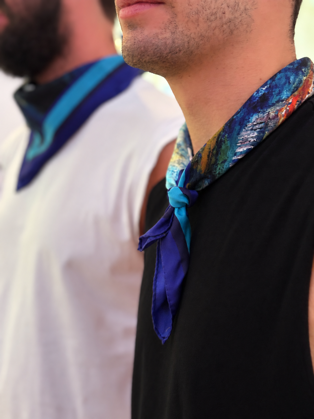 A close-up lifestyle shot of a man wearing a vibrant blue and multicolor patterned silk neck scarf tied around his collar over a black sleeveless shirt, with another man in a white shirt blurred in the background.