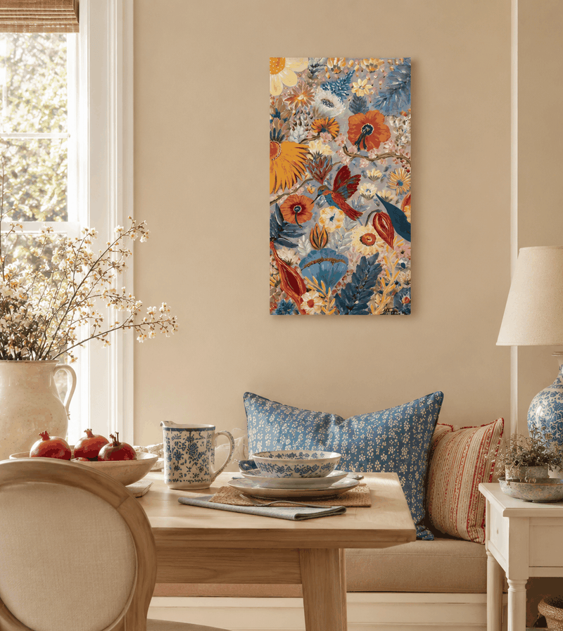 A colorful floral and bird canvas wall art print hangs on a beige wall in a styled dining room setting with a wooden table, blue patterned pillows, a lamp, and a window.