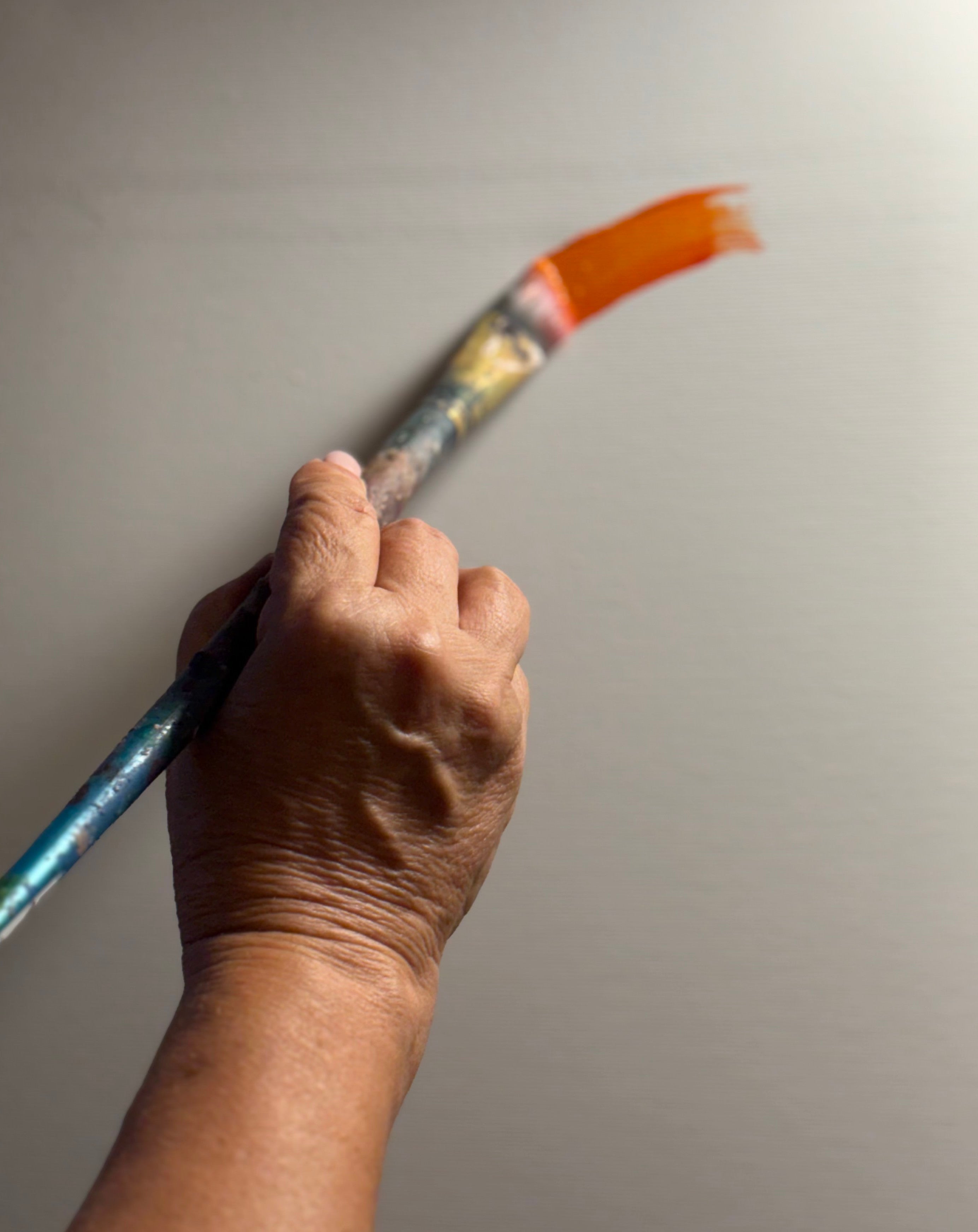 A close-up of a hand holding a paintbrush with orange bristles and a blue handle, pointing toward a plain light gray wall.