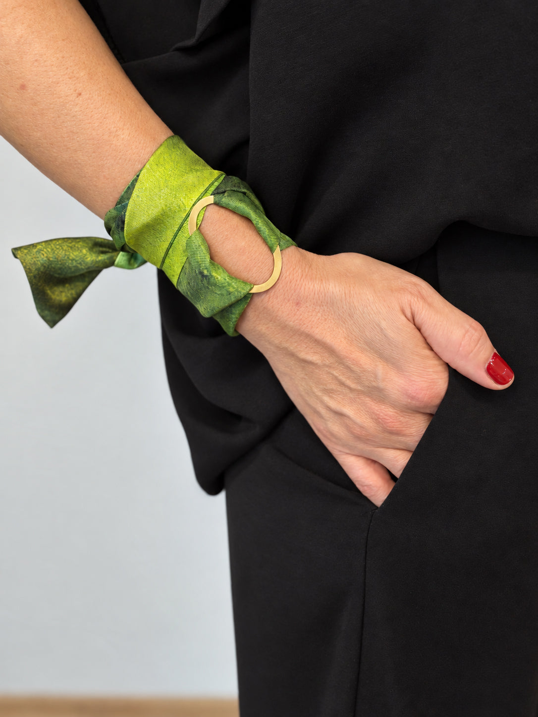 A close-up of a person's wrist wearing a green silk scarf bracelet with a gold metal ring cutout, paired with a black outfit.