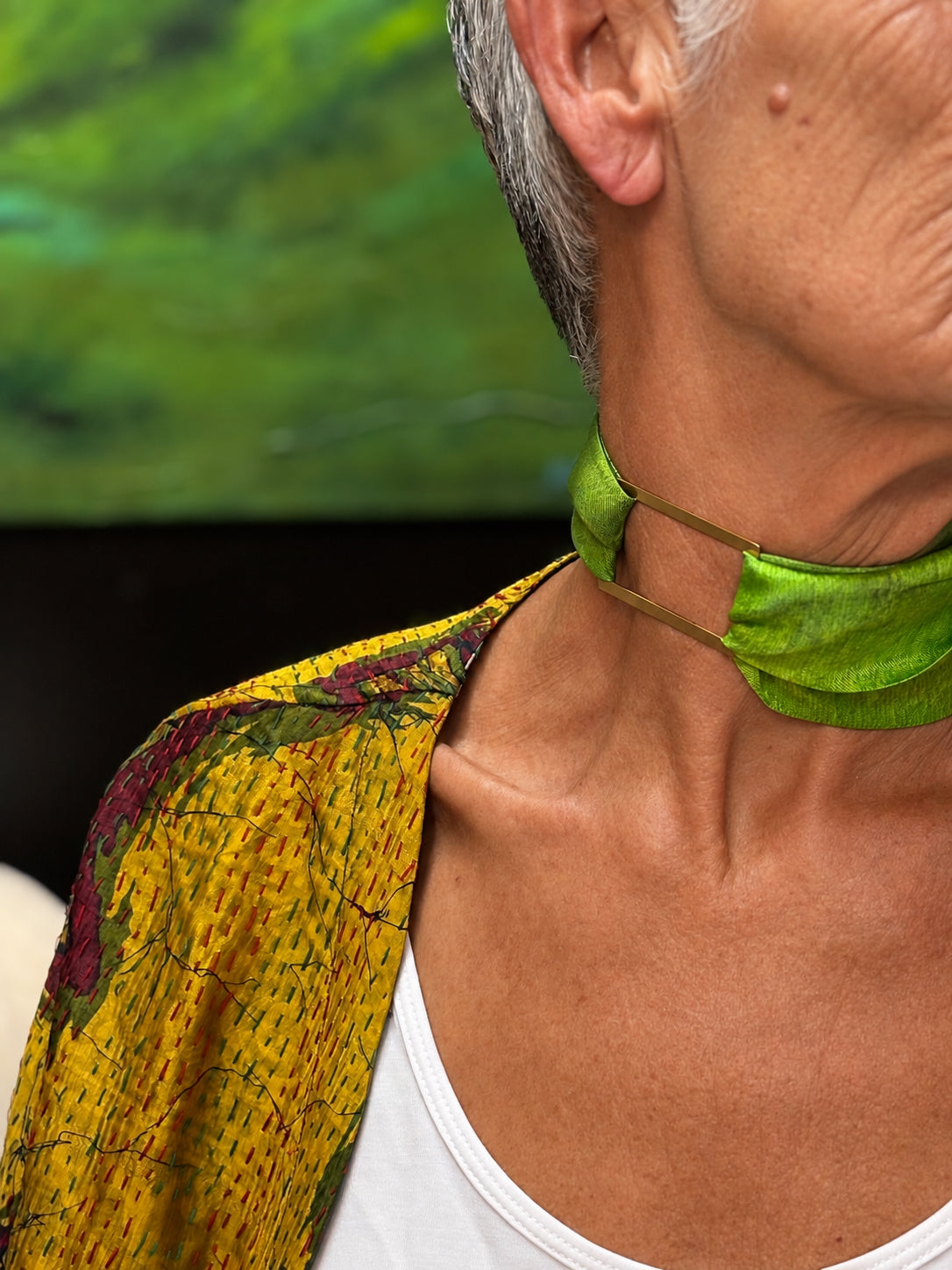 A close-up lifestyle shot of a person wearing a vibrant lime green fabric neck gaiter or face mask. The person has gray hair and is wearing a yellow patterned shawl over a white tank top, with a blurred green background.