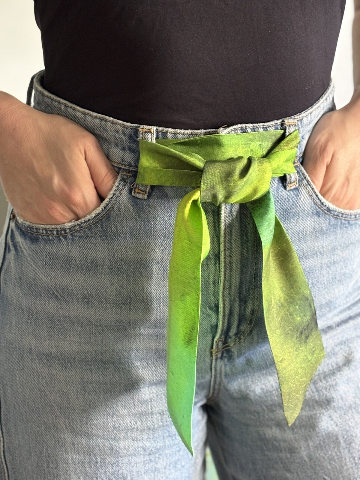 A close-up of a person wearing a bright green silk scarf tied around their waist as a belt, paired with light-wash blue jeans and a black top.