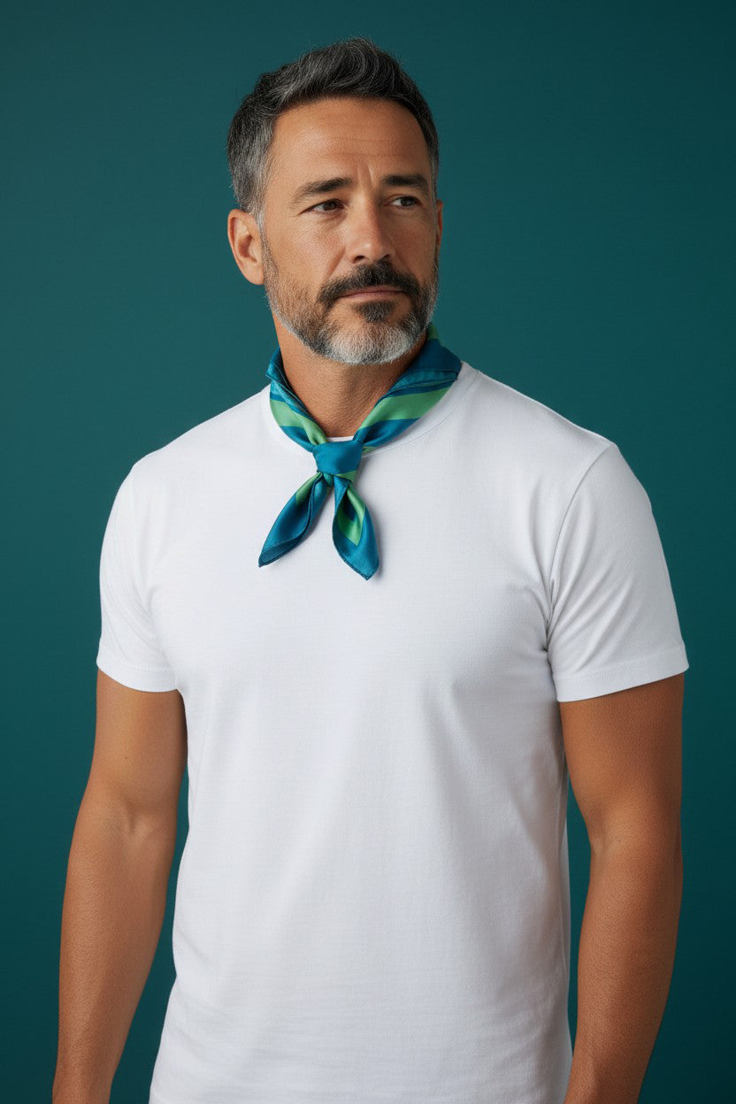 A man with a graying beard wearing a white t-shirt and a blue and green patterned silk neck scarf, standing against a solid dark teal background.