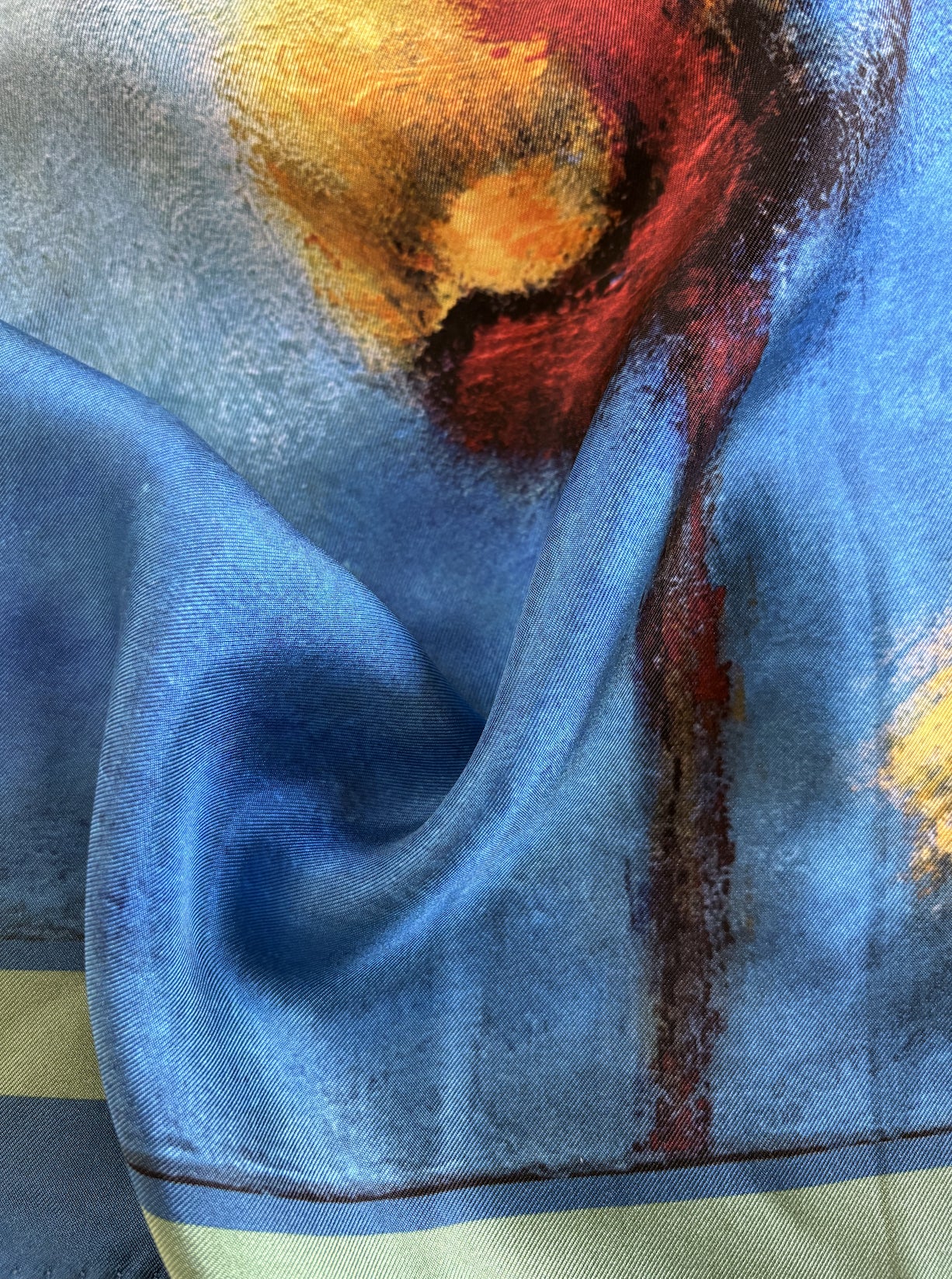 A close-up of a blue satin fabric with an abstract painted design in red, orange, yellow, and dark brown, draped to show its sheen and texture.