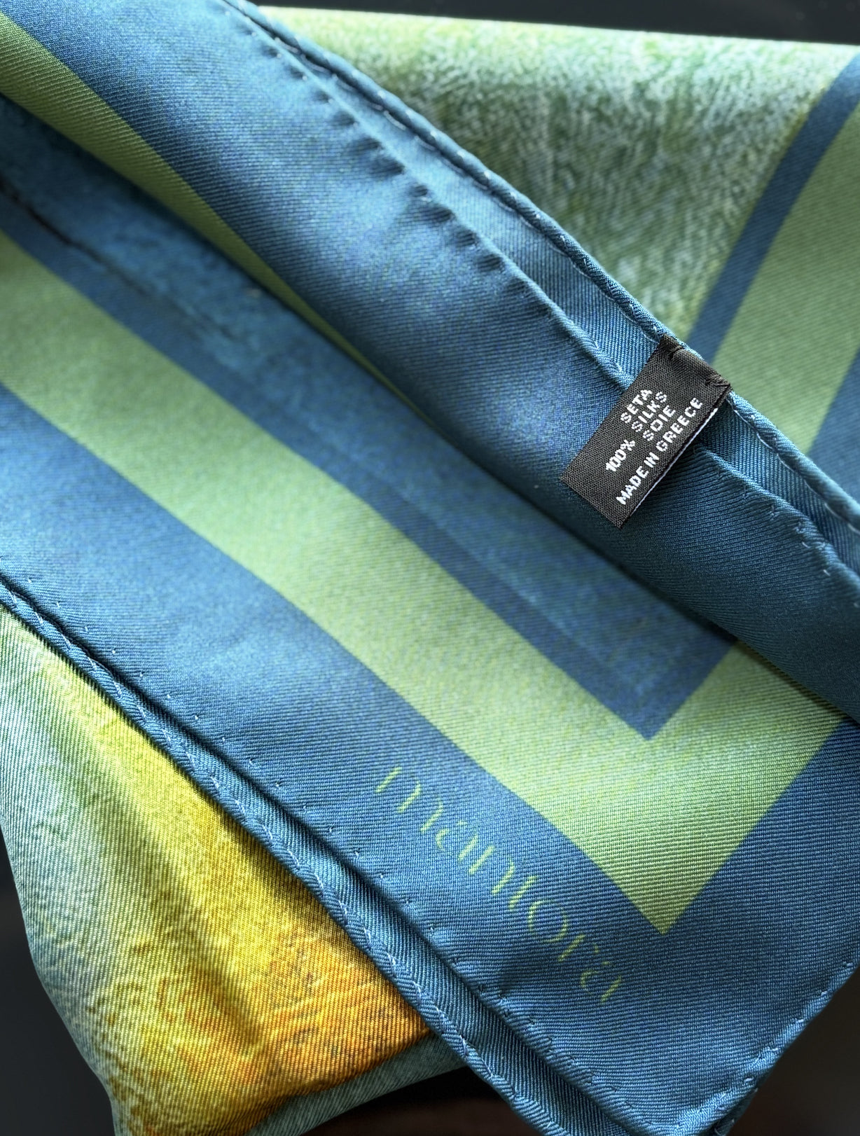 A close-up of a folded silk scarf featuring a blue border, a light green and yellow striped pattern, and a black brand label reading 'MANTOVA'.
