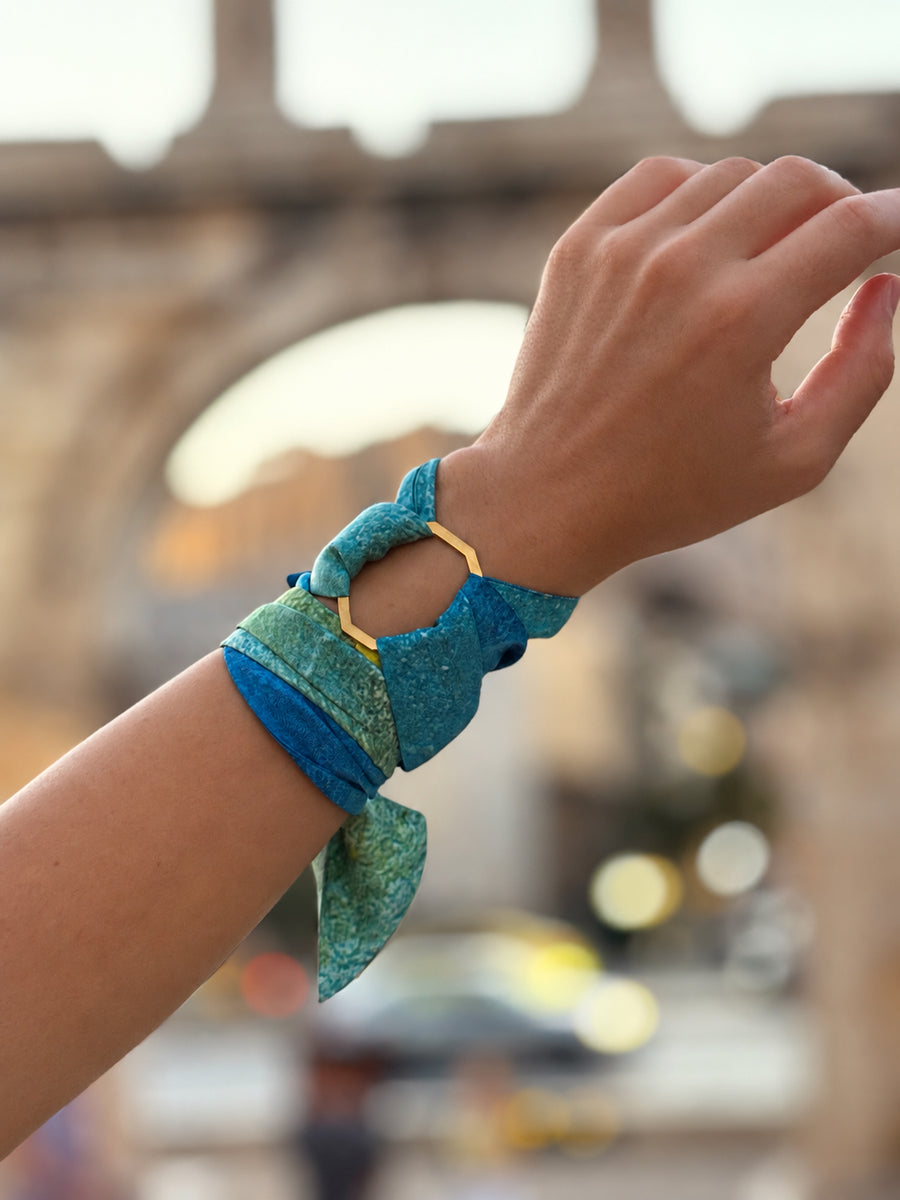 A close-up of a person's wrist wearing a blue and green patterned fabric bracelet with a gold-tone geometric ring, set against a blurred outdoor background.
