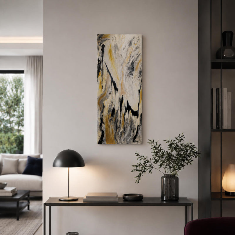 A vertical abstract canvas print with black, white, gray, and gold brushstrokes, displayed on a light gray wall in a modern living room setting above a console table with a lamp and a plant.