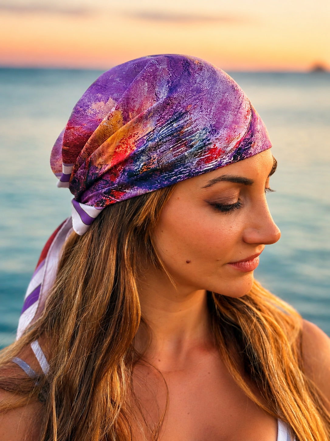 A woman in profile wearing a colorful abstract print headscarf with purple, pink, and blue tones, set against a soft-focus ocean sunset background.