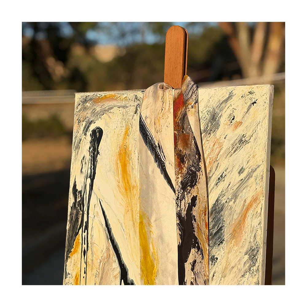 An abstract canvas painting with cream, black, gray, and ochre yellow brushstrokes, displayed on an easel outdoors with a blurred natural background.