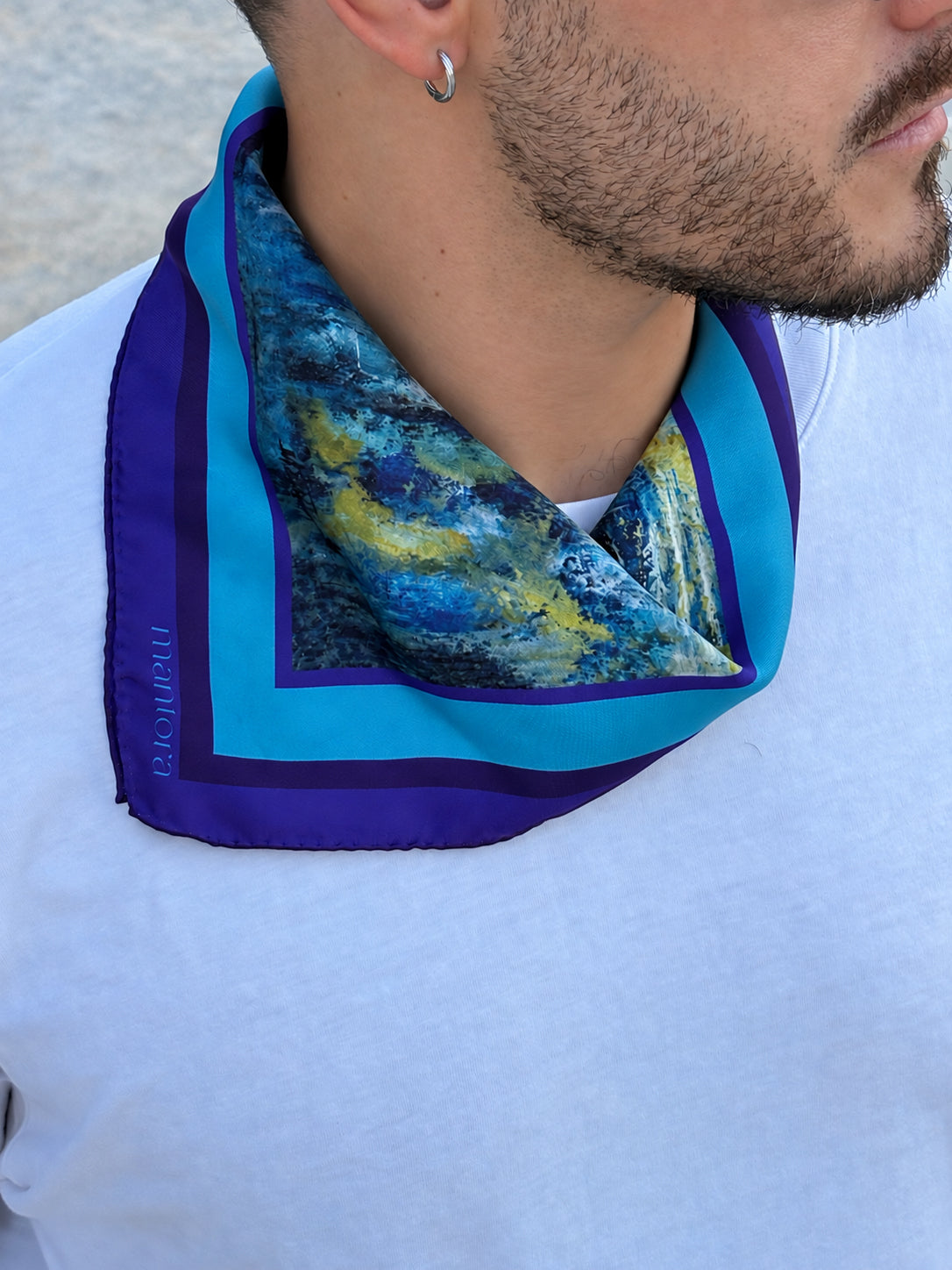 A close-up of a man wearing a square silk scarf with a blue and yellow abstract print, featuring a purple border and turquoise trim, styled around his neck over a white t-shirt.