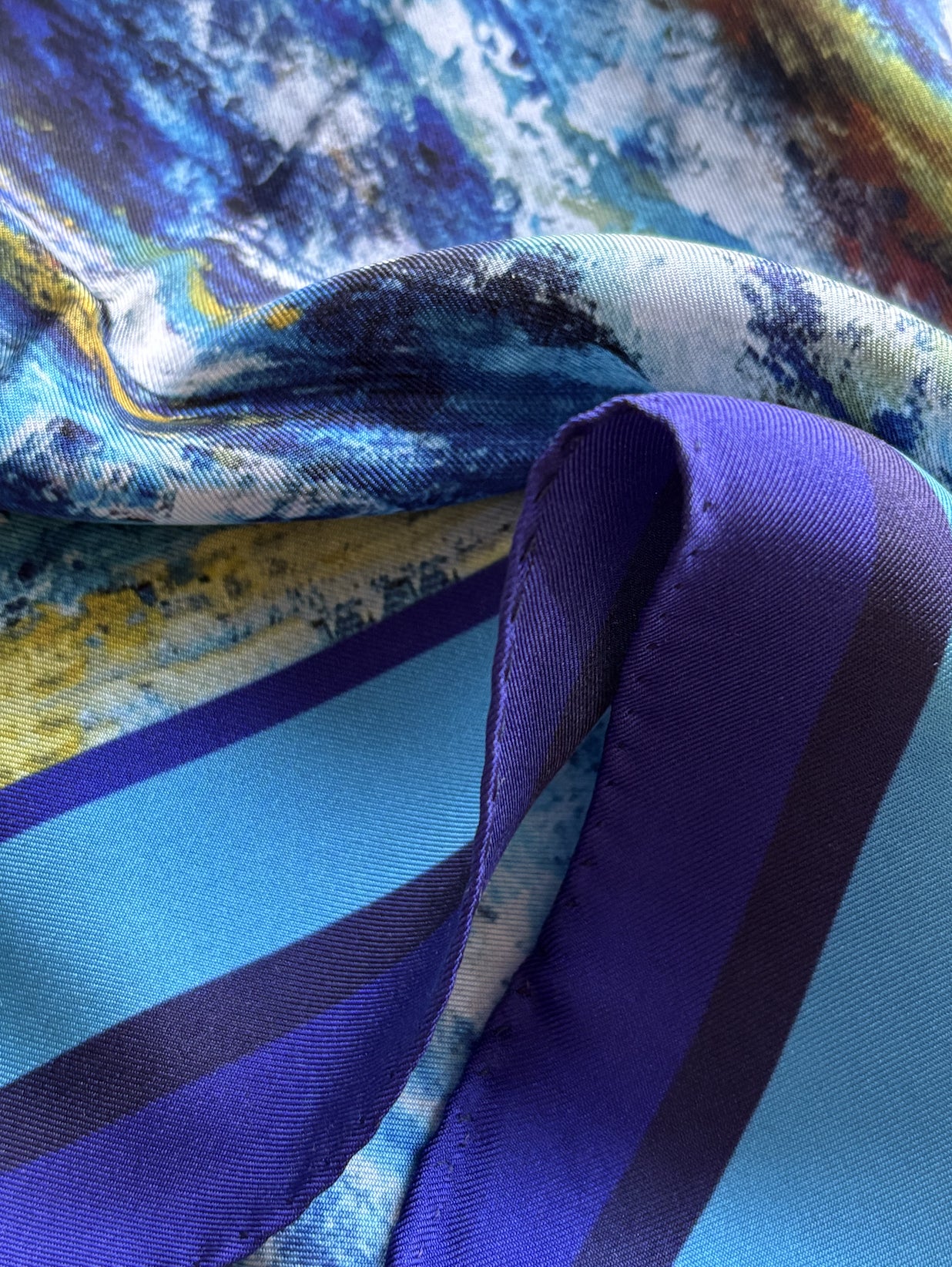 A close-up of a folded silk scarf featuring a blue and purple abstract landscape print with a solid purple border.