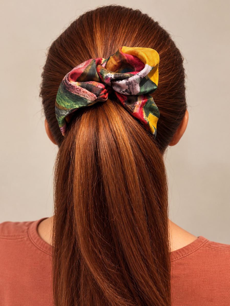 A person shown from the back wearing a brown hair extension or wig with a large multicolor fabric scrunchie at the top, paired with a terracotta-colored top against a plain beige background.