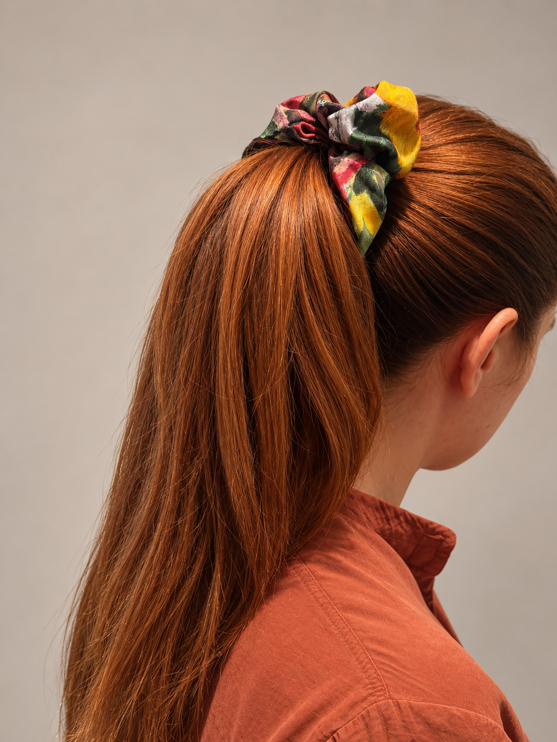 A person with long auburn hair wearing a colorful patterned fabric scrunchie, shown from the side against a plain light gray background.