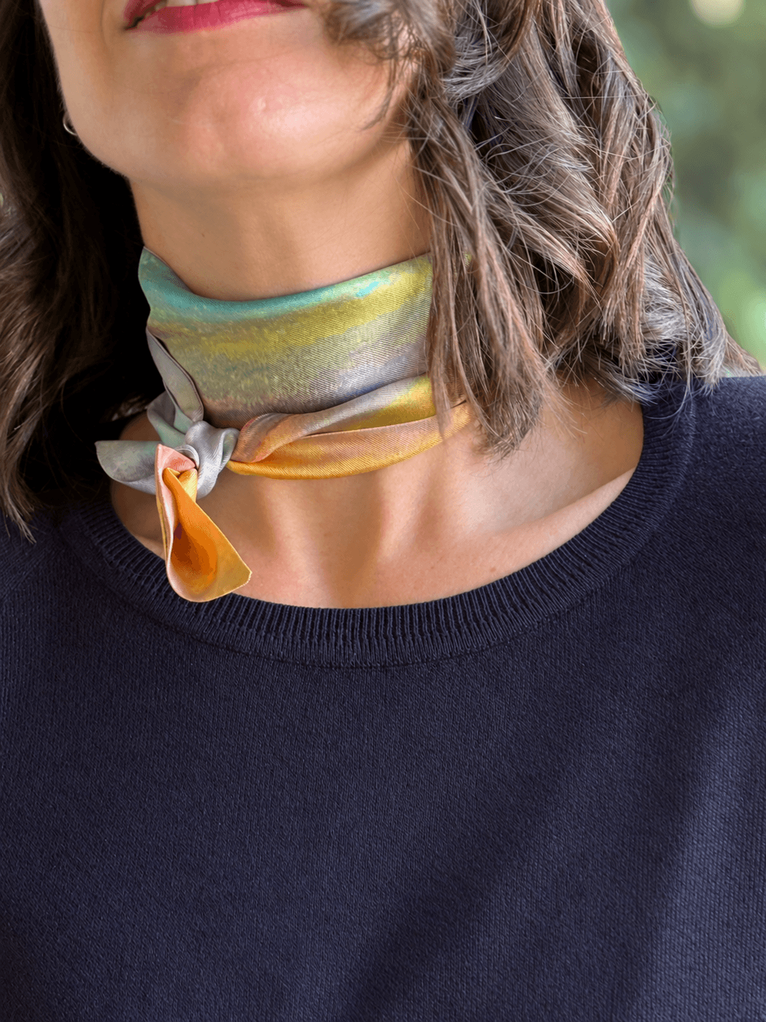 A close-up of a woman wearing a silky scarf tied around her neck. The scarf has an iridescent, metallic finish shifting between yellow, green, blue, and orange-pink, paired with a dark navy knit top.