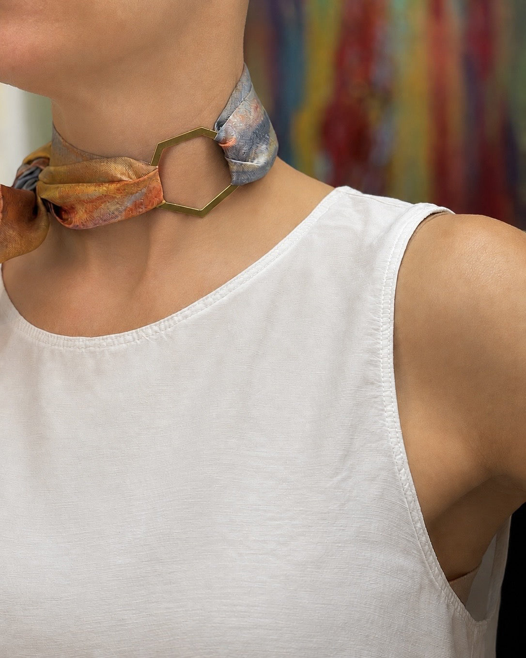 A close-up of a person wearing a white sleeveless top and a patterned scarf styled as a choker necklace with a geometric metal ring.