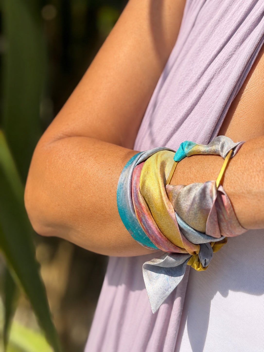 A close-up of a person's forearm wearing multiple satin-like fabric bracelets in pastel colors, including turquoise, pink, yellow, lavender, and gold, against a softly blurred outdoor background.