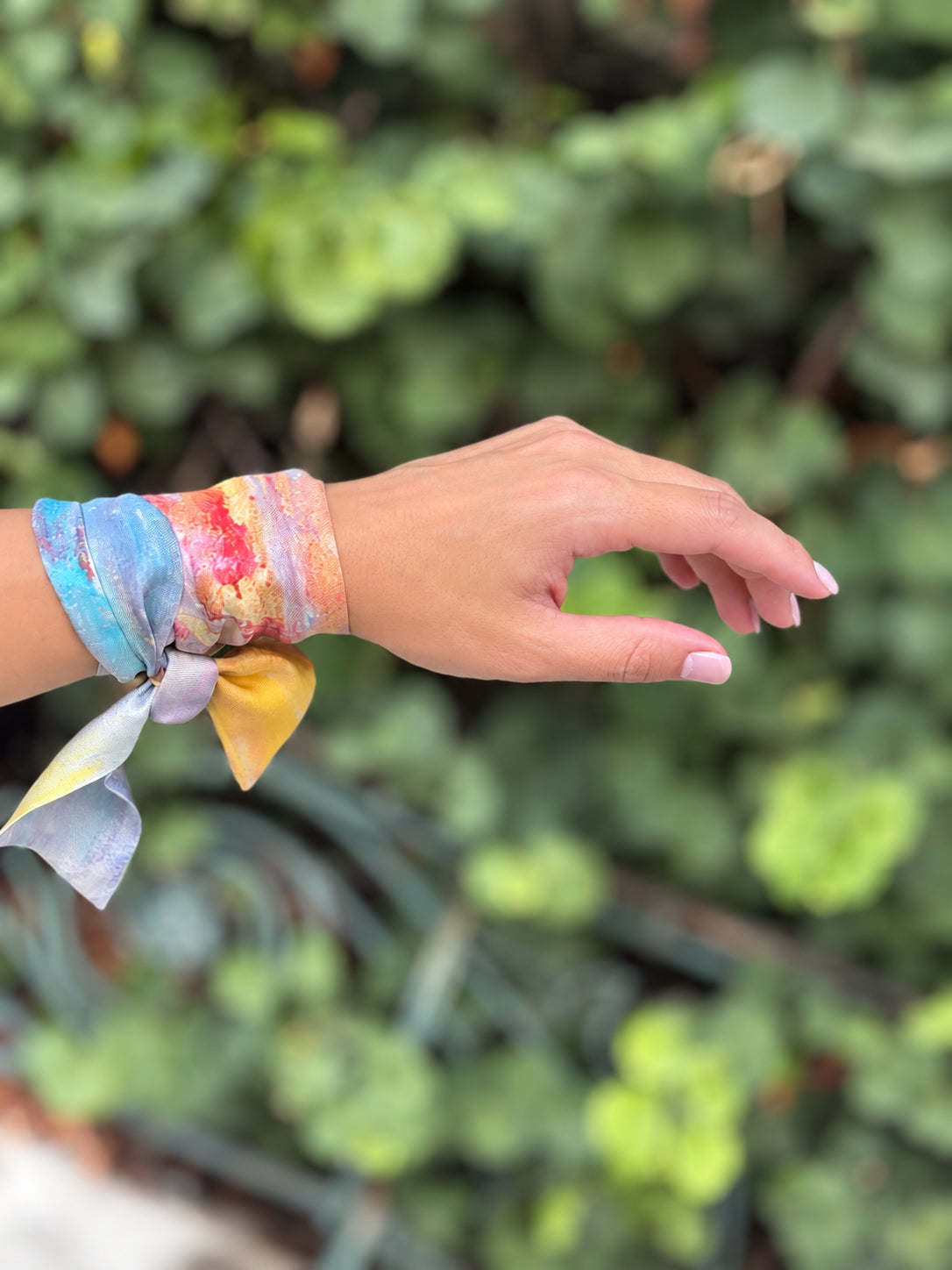 A multicolor tie-dye silk scarf wrapped around a person's wrist, shown against a blurred green garden background.
