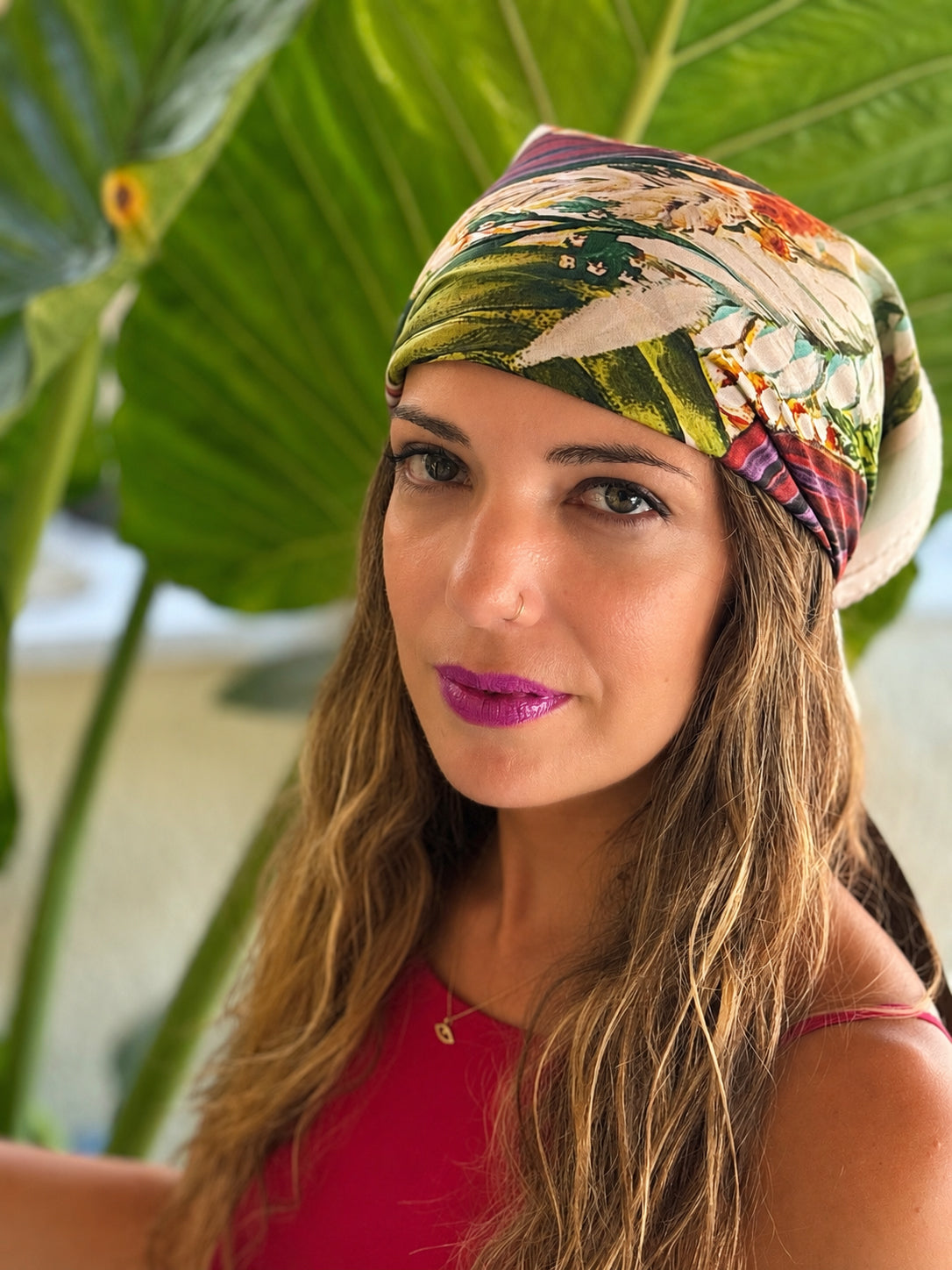 A woman wearing a colorful tropical print headscarf, photographed in a lifestyle setting with large green tropical leaves in the background.