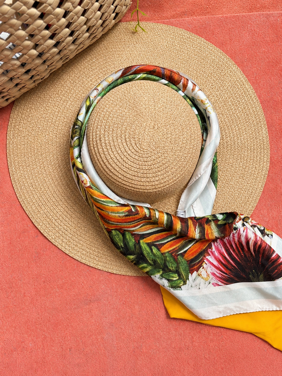 A straw sun hat with a colorful tropical print scarf draped over it, styled on a terracotta surface next to a woven basket.