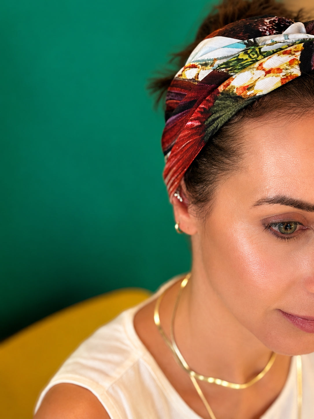 A close-up of a woman wearing a colorful, patterned fabric headband with a swirl design, paired with gold necklaces and earrings, set against a solid green background.