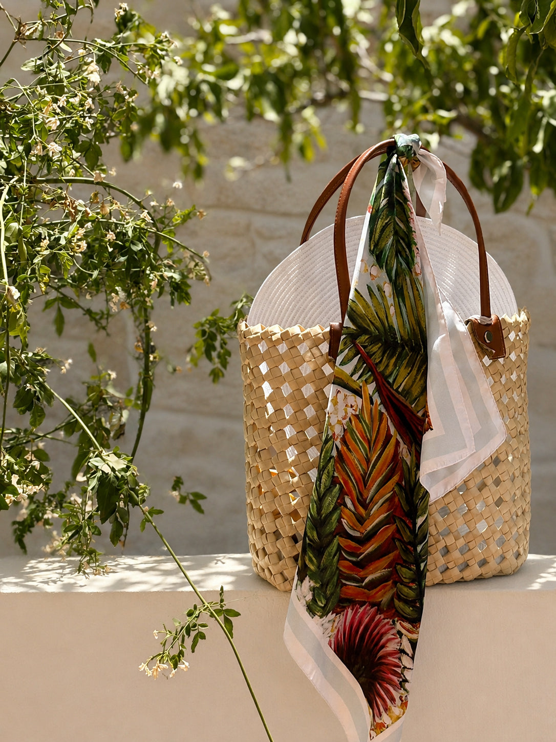 A woven straw tote bag with brown leather handles, draped with a tropical palm leaf print scarf, set against a background of green foliage and a light stone wall.