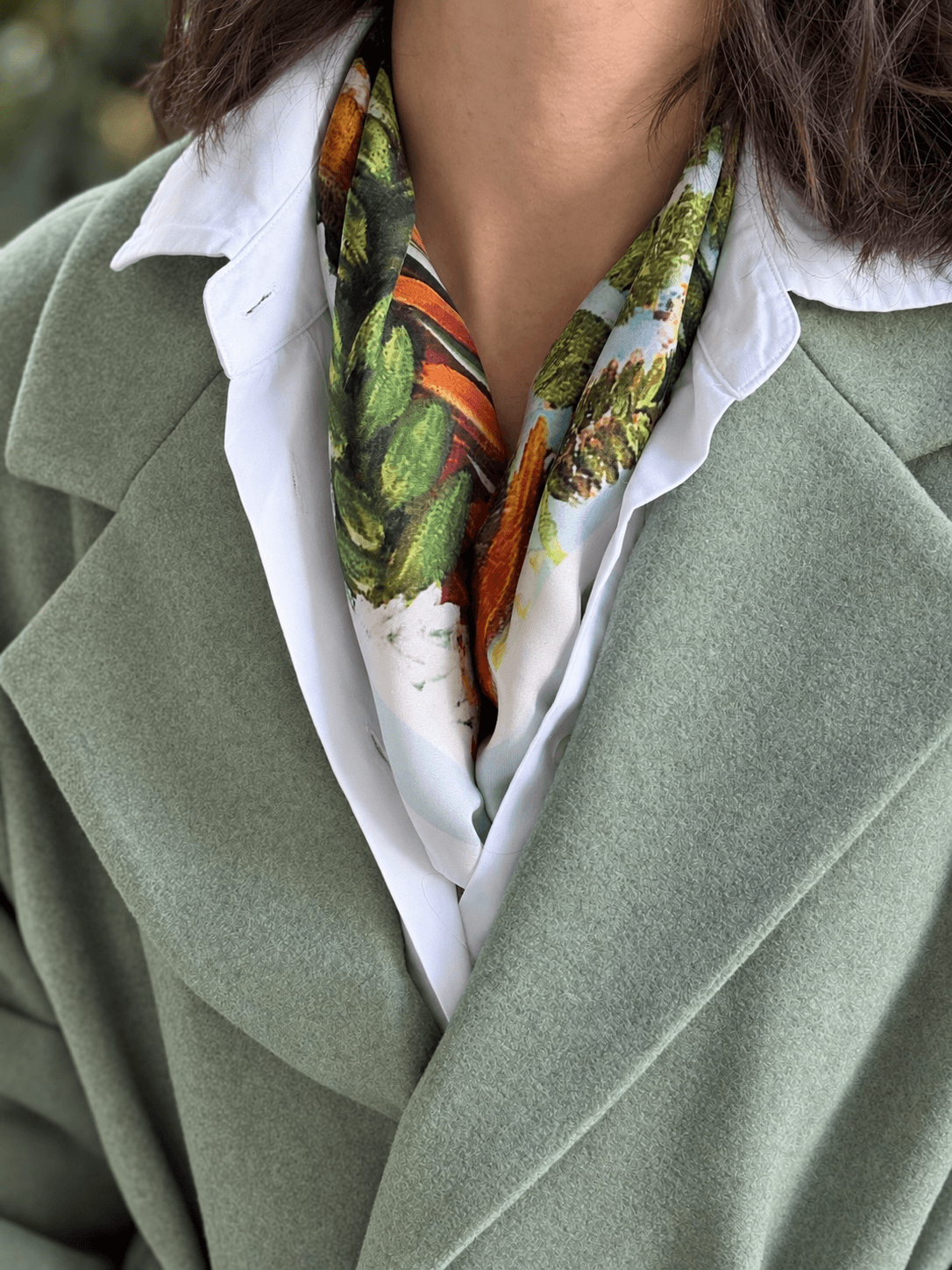 A close-up of a person wearing a sage green wool coat over a white collared shirt, with a colorful botanical silk scarf tied around the neck.