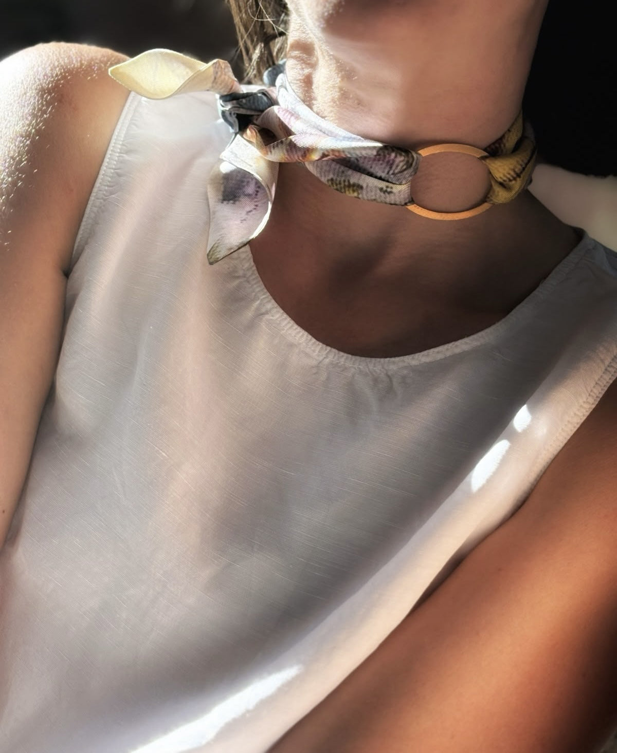 A close-up lifestyle shot of a model wearing a cream-colored sleeveless top and a patterned silk neck scarf tied around a gold ring choker.