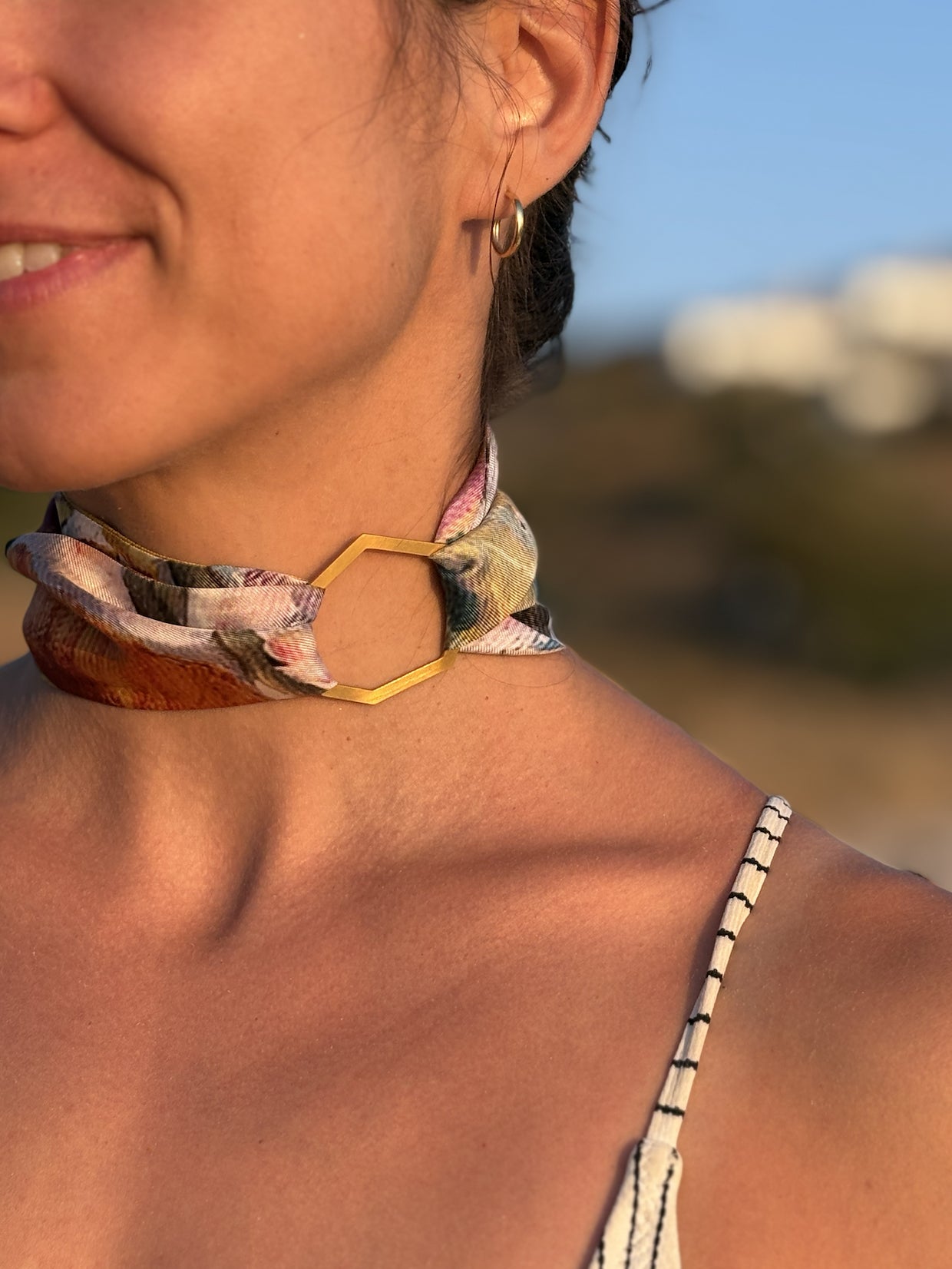 Close-up of a woman outdoors wearing a patterned scarf choker with a gold hexagonal metal connector and a small gold hoop earring.