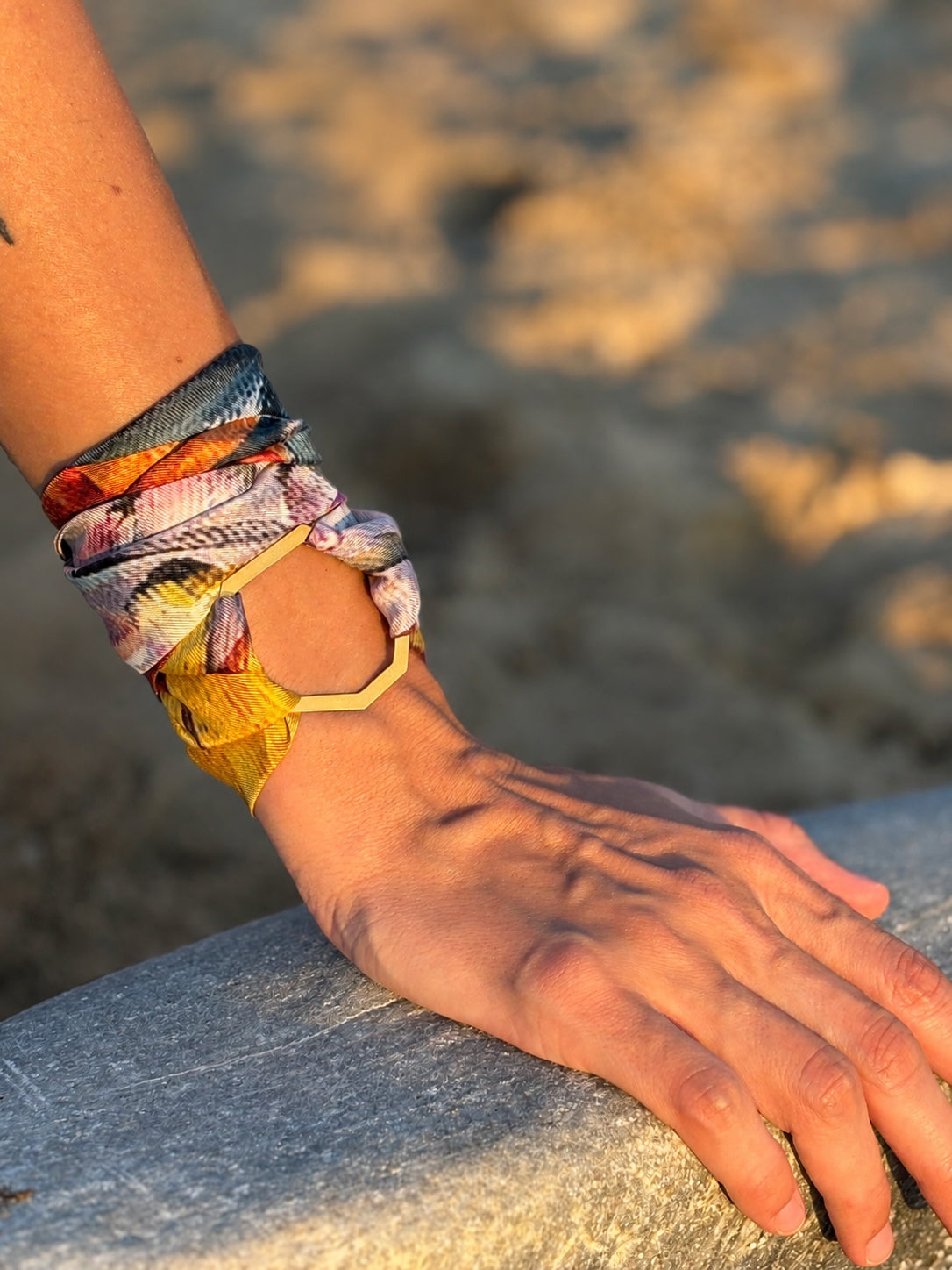A close-up of a person's wrist and hand wearing a colorful patterned wrap bracelet with a gold geometric accent, resting on a stone surface outdoors.