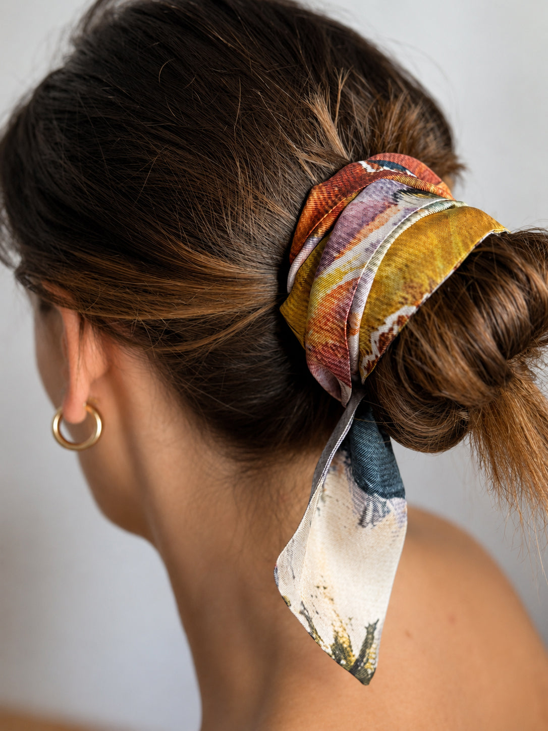 A close-up side view of a woman's head with her hair in a low bun, secured with a patterned silk scrunchie. The scrunchie features an abstract painterly design in orange, yellow, blue, and white.