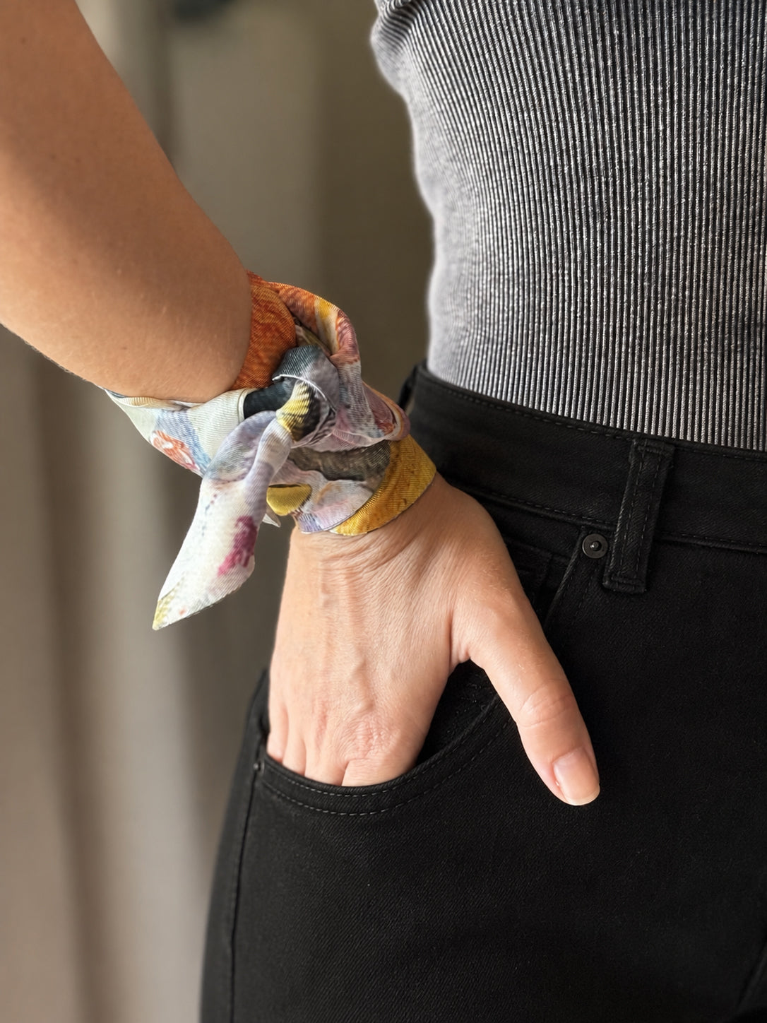 A close-up of a person wearing a multicolor floral silk scarf as a wrist tie, paired with a gray ribbed top and black pants.