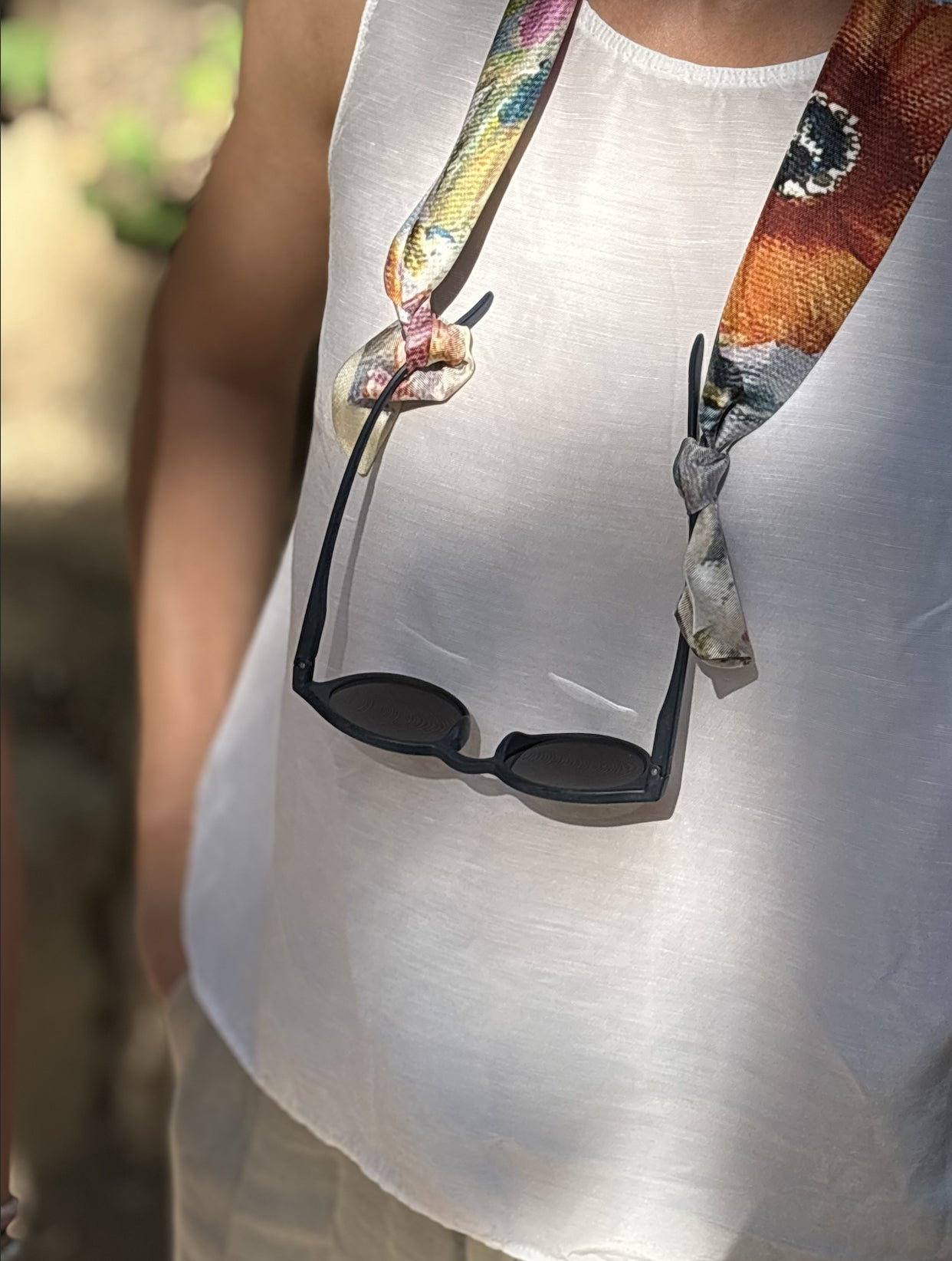 A close-up lifestyle shot of a person wearing a white tank top and beige shorts, with black sunglasses hanging from a colorful patterned neck strap tied around their neck.