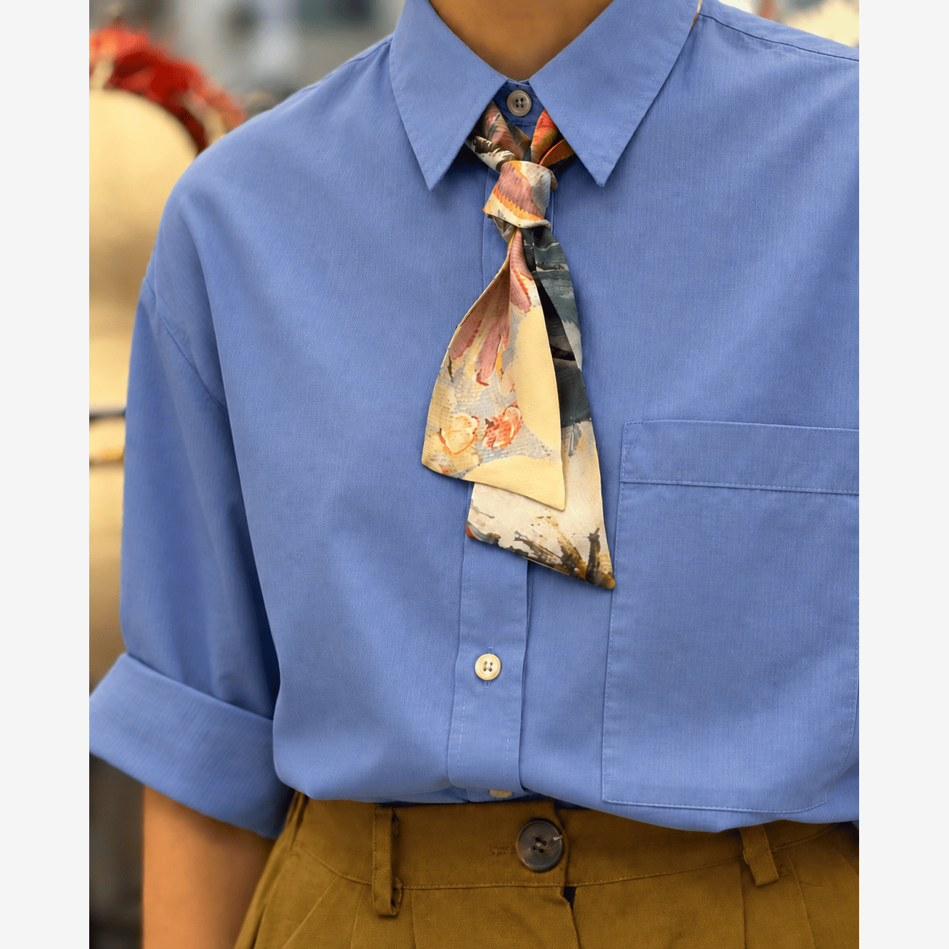 A close-up of a person wearing a light blue short-sleeve button-up shirt with a patterned necktie and brown trousers. The necktie has a beige base with orange, red, and dark gray abstract print details.