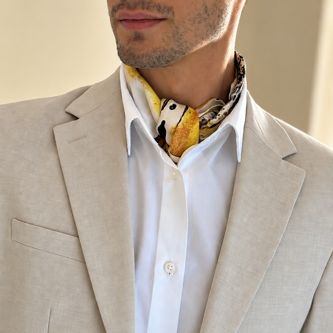 A close-up of a man wearing a light beige linen blazer, a white button-down shirt, and a patterned yellow, black, and white neck scarf.
