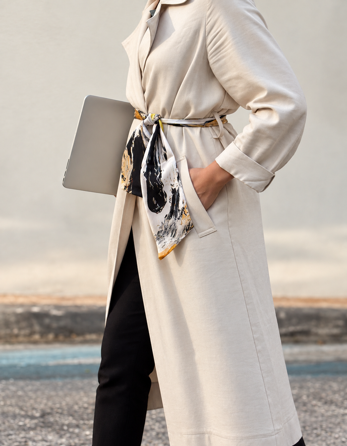 A woman wearing a long cream trench coat with a matching belt and a patterned silk scarf, holding a gray laptop, standing outdoors against a soft-focus background.