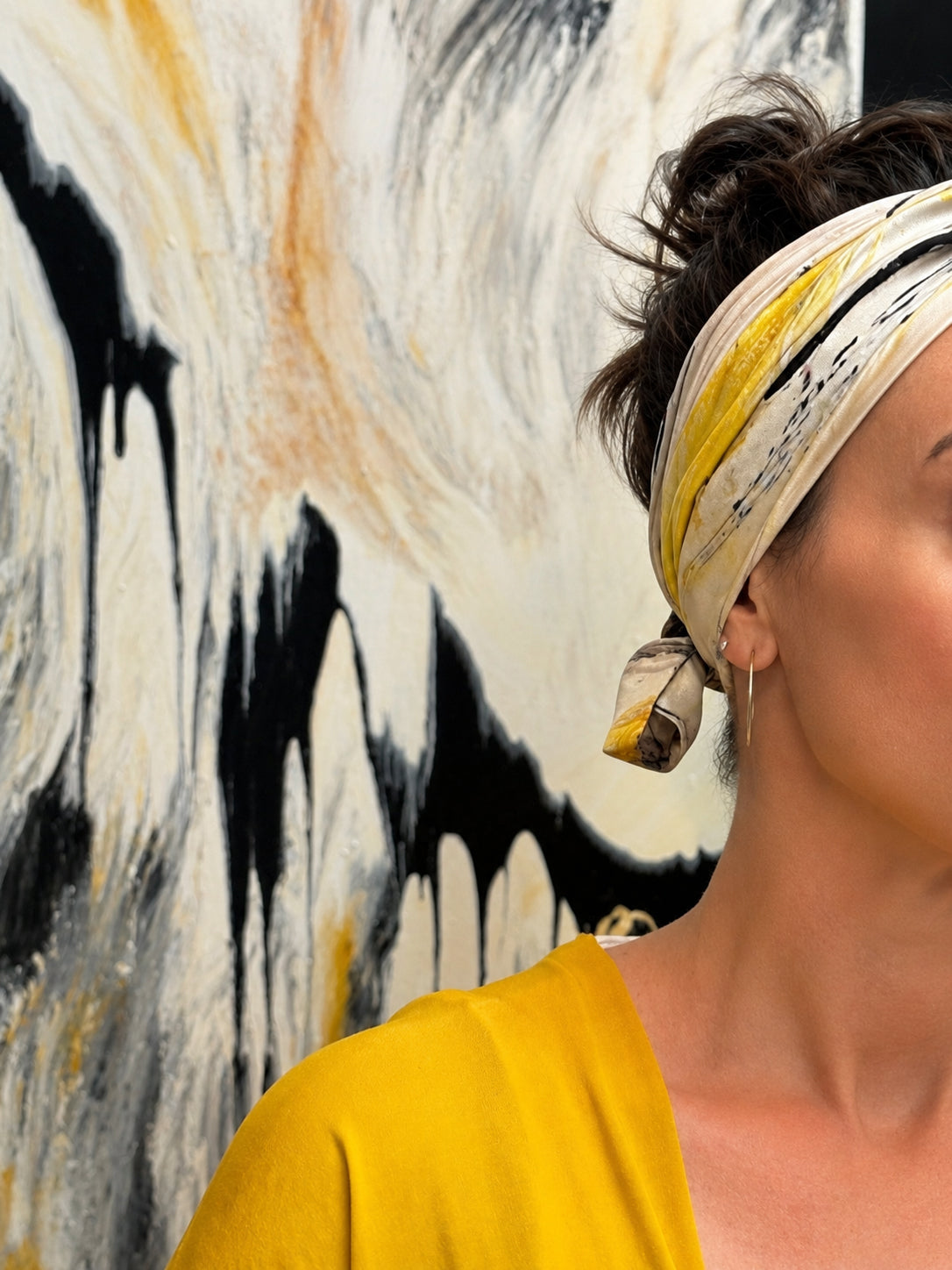 A close-up side profile of a woman wearing a yellow and white patterned silk headband, standing in front of an abstract black, white, and yellow painting.