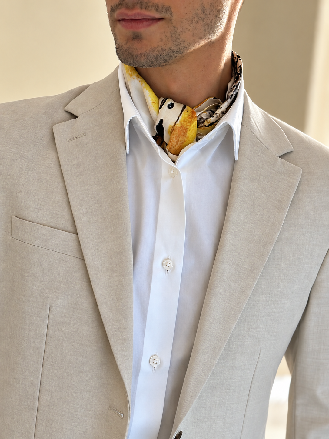 A close-up of a man wearing a light beige linen suit jacket over a white button-down shirt, accessorized with a yellow and black patterned neck scarf.
