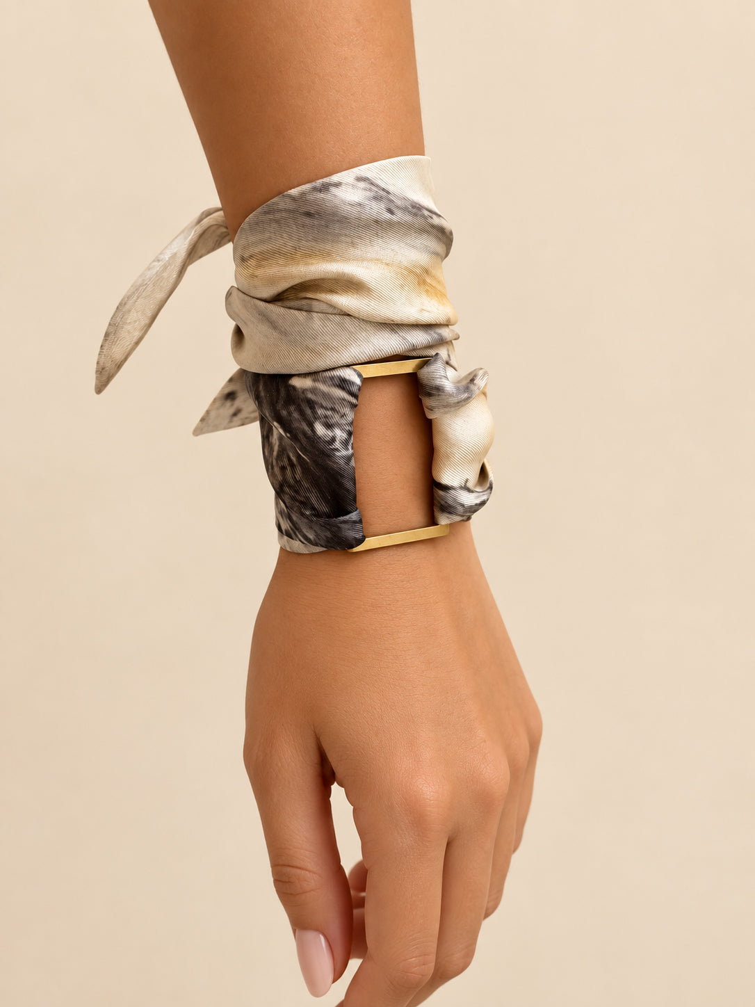 A close-up of a person's wrist and hand wearing a gold-tone cuff bracelet wrapped with a patterned silk scarf in beige, gray, and black tones, set against a solid beige background.