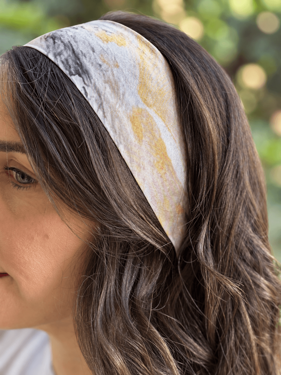 A close-up side profile of a woman wearing a wide fabric headband with a marbled pattern in white, gray, and muted gold, set against a soft-focus green outdoor background.