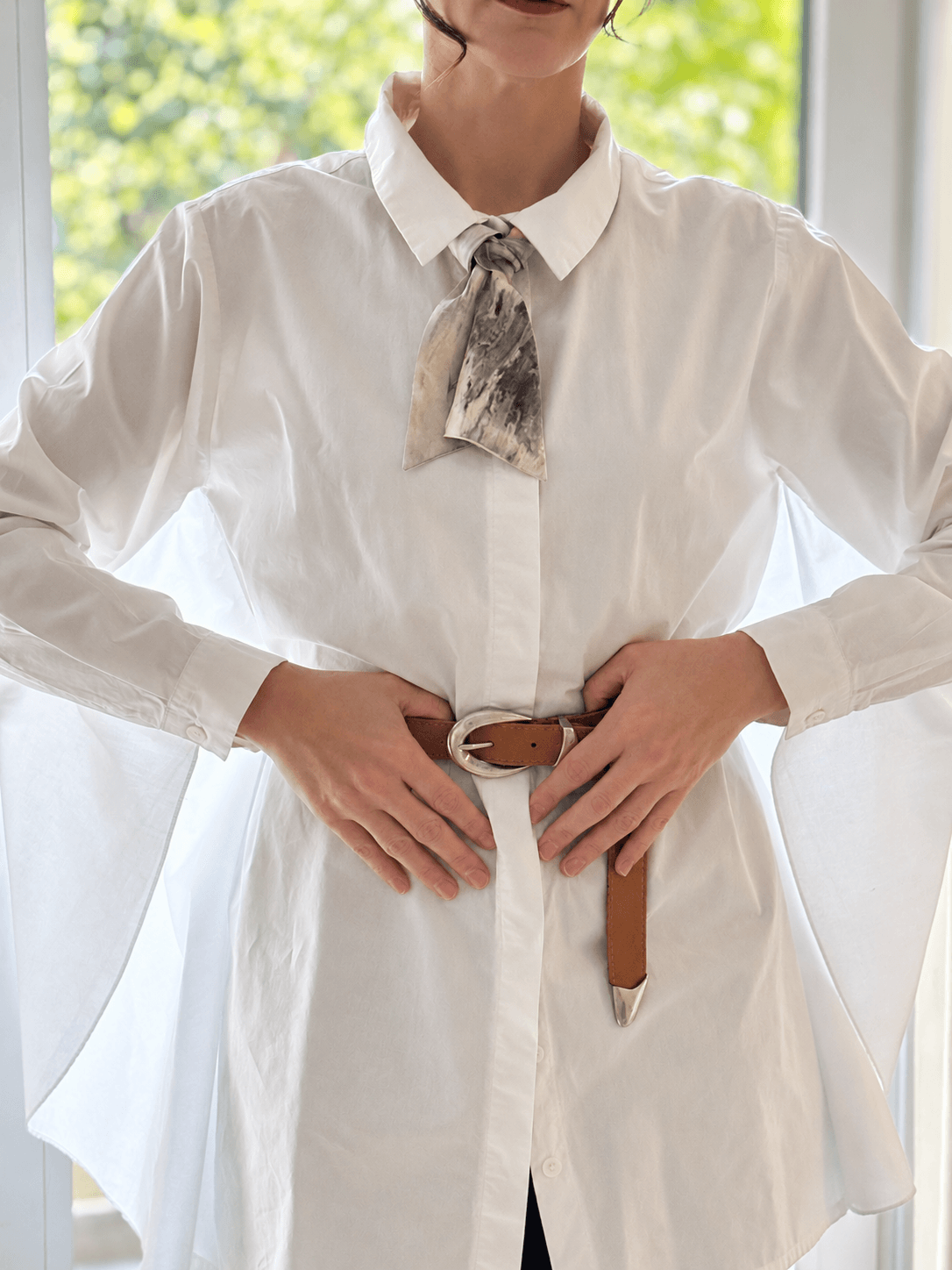 A woman wearing a white long-sleeve button-up shirt with a patterned neck scarf and a brown belt, standing in front of a bright window.