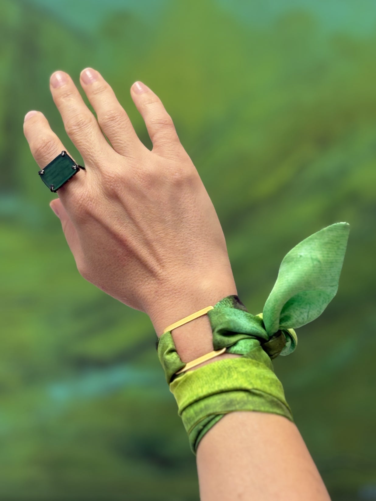 A close-up of a person's hand and wrist wearing a green rectangular ring, a green silk scarf tied into a knot, and a green fabric bracelet with a gold buckle, set against a blurred green background.