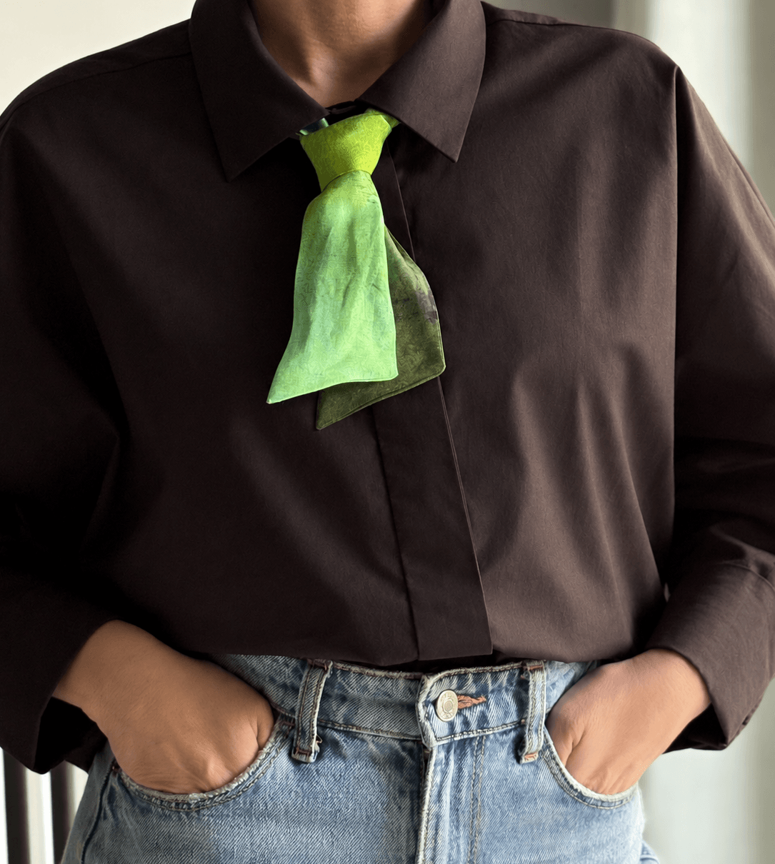 A person wearing a dark brown long-sleeve button-up shirt with a bright green scarf tied around the collar, paired with light blue jeans.