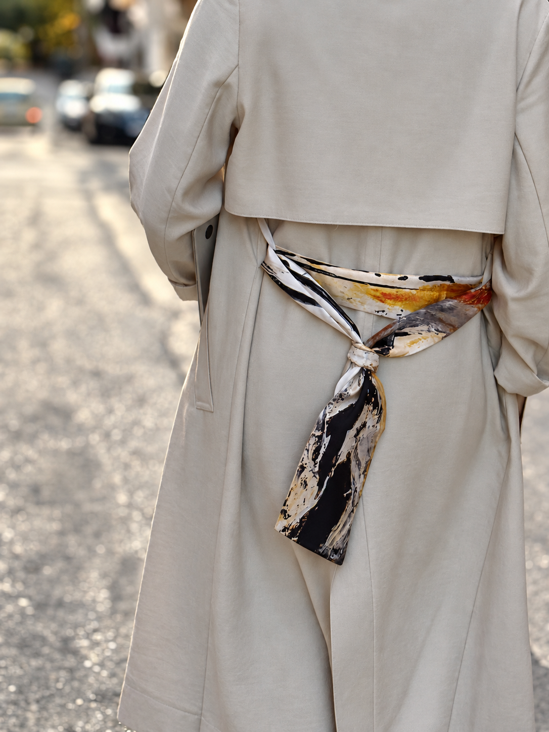 A close-up back view of a person wearing a light beige trench coat and wide-leg trousers, tied at the waist with a patterned scarf in black, beige, and white, set against a blurred street background.